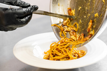 Chef plating creamy pasta with bacon using tongs in a professional kitchen. Close-up of food preparation, showcasing culinary technique and gourmet presentation.