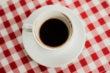 Top view of a white cup filled with black coffee placed on a red and white checkered tablecloth, minimalist breakfast concept.