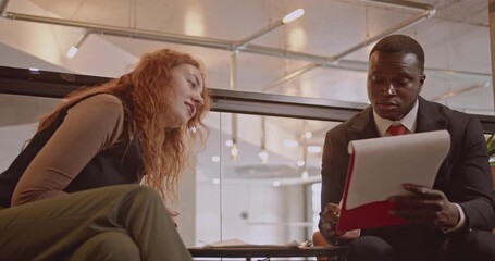 Two diverse business colleagues discuss a project together in their modern office space. They review documents and engage in professional conversation. - Powered by Adobe