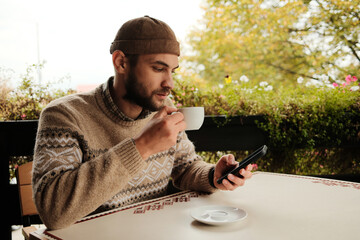 Young man in brown sweater holding coffee cup and using smartphone at outdoor cafe table, casual modern lifestyle scene