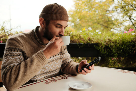 Young man in brown sweater holding coffee cup and using smartphone at outdoor cafe table, casual modern lifestyle scene - Powered by Adobe