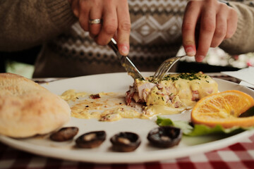 Close-up of hands cutting grilled meat dish topped with creamy cheese sauce and garnished with herbs, bread served with mushrooms and orange slice