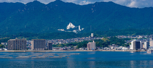Calm blue waters with oyster rafts sit before Hiroshima, with apartments by the shore and a white hilltop complex, looking toward Miyajima on a clear day.