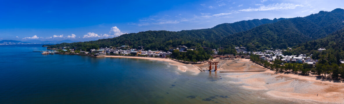 Aerial daytime view of the Great Torii at Itsukushima Shrine on Miyajima, Hiroshima, Japan, in shallow tidal water, with Seto Inland Sea, beach, town, and forested hills.