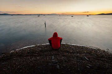 Person in red hoodie sitting by the sea at sunset
