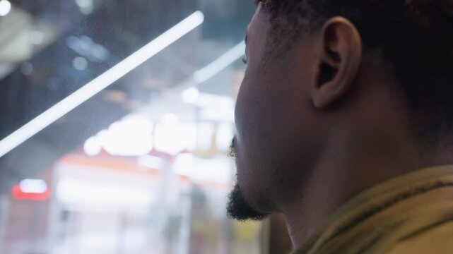 Close up of young artist with braided hairstyle and trimmed beard standing inside ascending elevator, gazing thoughtfully through glass wall as bright reflections and blurred shapes