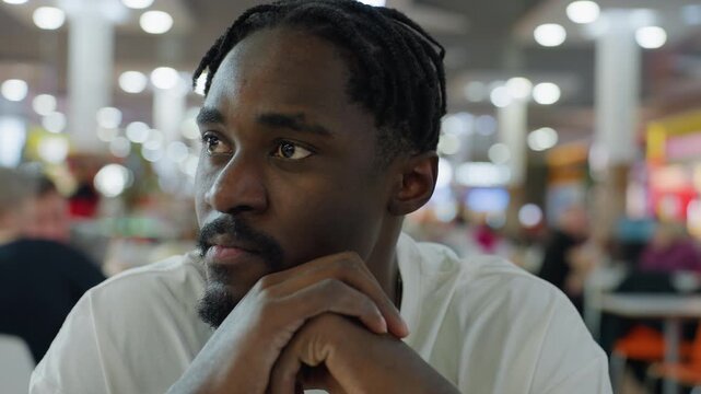 Young african man in white shirt sitting with hands clasped under chin looks sideways observing people in crowded modern restaurant with blurred lights and diners in colorful indoor environment