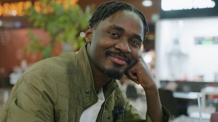 Close up of young man posing with warm smile resting hand under chin, wearing green jacket in indoor setting with blurred background lights, relaxed thoughtful mood