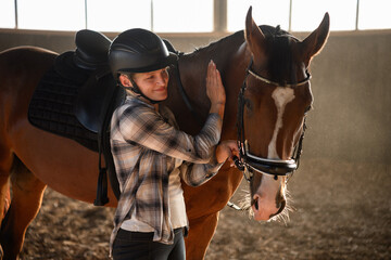 Female jockey wearing riding helmet stands in front of horse in arena, stroking it with hand and...