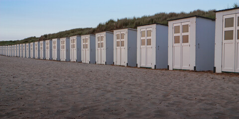 A panoramic shot of a long, neat line of small white beach cabins backed by a high grassy dune on the sandy shore of Texel at dusk.