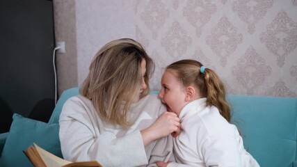 Mother and daughter sitting on sofa sharing joyful playful moment while reading book together, smiling and looking at each other with love, family bonding time, warmth, comfort, happiness, connection