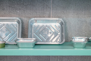 Multiple sizes of disposable aluminum food containers displayed neatly on a turquoise shelf with a gray textured wall in the background