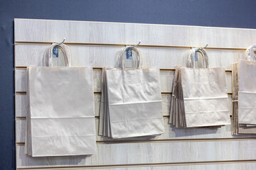 Plain paper shopping bags hang on metal hooks on a wooden panel on a store wall. Packaging of goods