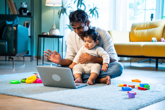 Indian Father and infant boy share laptop video call while sitting on carpet in modern home