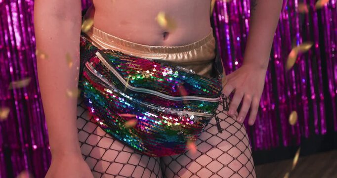 Carnival party, waist close-up on dancer in gold shorts, fishnet stockings, multicolor sequin fanny pack. Golden confetti flies with hipsway, festive energy builds to radiant smile and purple makeup.