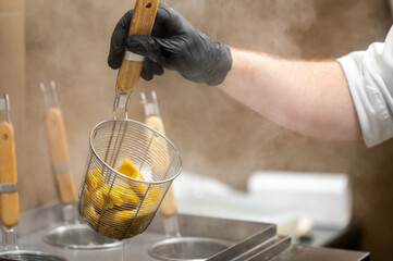 Chef using metal basket to remove cooked ravioli from boiling water in commercial kitchen. Steam rises, glove worn for safety. Multiple baskets suggest high-volume pasta preparation.