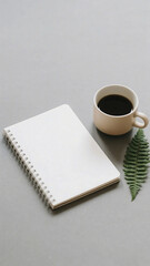 A notebook, a cup of coffee, and a fern leaf on a gray surface, creating a simple and peaceful scene.