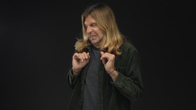 Blond man with long hair scrunches face and pinches fingers near chest in black studio set; disgust.