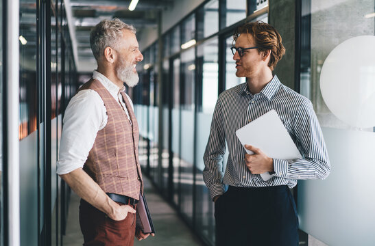 Senior man explains project visuals on glass board while young male colleague with digital tablet listens attentively. Scene illustrates visual strategy and tech-enabled team alignment.