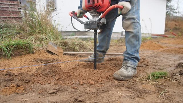 A construction worker drills a hole in the ground at a construction site with a manual drilling rig for pouring concrete