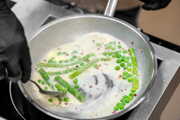 Chef stirs creamy vegetable sauce with peas and asparagus in hot pan. Steam rises during cooking. Close-up of professional kitchen preparation.