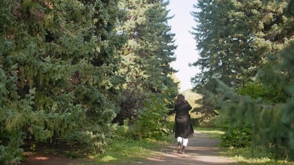 long shot nature lover in long winter coat strolling through arboretum, holding phone and raising it to take shots among tall pines, peaceful walk capturing greenery during relaxed autumn day