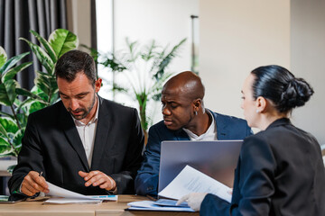 Group of three professionals engaged in a business meeting, discussing documents and strategies, surrounded by greenery in a modern office environment, showcasing collaboration and teamwork