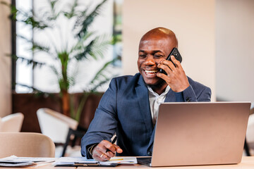 African American man in a business suit is engaged in a phone conversation while working on a...