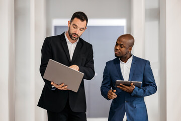 Two business professionals engaged in discussion, one holding a laptop and the other a tablet, in a modern office environment showcasing collaboration and teamwork