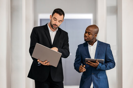 Two business professionals engaged in discussion, one holding a laptop and the other a tablet, in a modern office environment showcasing collaboration and teamwork