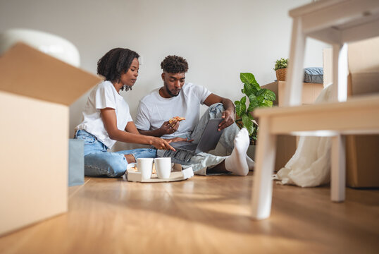 Smiling African American couple sitting on the floor among moving boxes, using a laptop together. Happy moment of love, teamwork, and excitement while planning their new home life