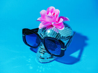 Party disco ball style scull with pink rose and large blue sunglasses on blue color background. Extravagant person concept.