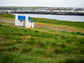 Blue and white benches on a grass with view on ocean and white houses on other side of bay. Stunning nature scenery in Kilkee town, county Clare, Ireland. Cloudy sky. Travel and tourism.