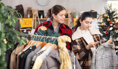 Two women stand in a store and choose outerwear, among jackets, coats and windbreakers against the background of a Christmas tree. Female friends buy stylish clothes on New Year sale