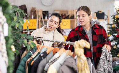 Two women stand in a store and choose outerwear, among jackets, coats and windbreakers against the background of a Christmas tree. Female friends buy stylish clothes on New Year sale