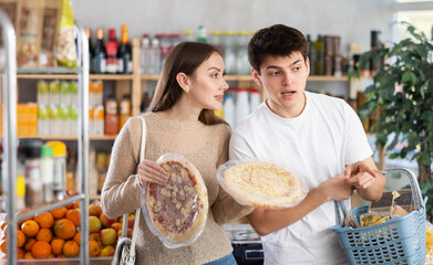 Happy young wife and husband choose large and fresh pizza in big supermarket in department of semi-finished products