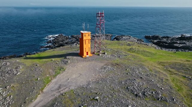 Aerial approach to lighthouse Hvalnes Iceland. Drone moves toward the orange tower surrounded by rocky shoreline and sea breeze.