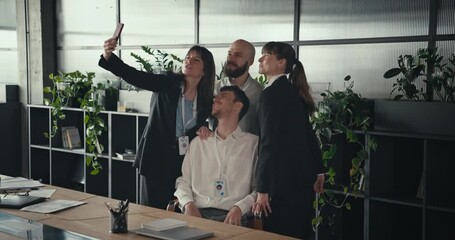 Four happy coworkers smiling and taking a selfie with a smartphone in a modern office with plants and shelves wearing ID badges and business attire