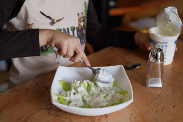 Boy adds salt to green lettuce salad at home. Concept of cooking habit, nutrition, and family lifestyle.
