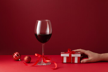 Elegant still life composition with a glass of red wine, Christmas ornaments, and a striped gift box in woman's hand on a deep red background
