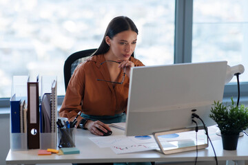 Concentrated lady manager or CEO sitting in front of computer monitor, working on marketing report at modern office, free space