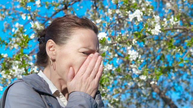 Woman scratching nose by flowers. view of unhappy woman sneezing and rubbing her nose during allergy attack in the spring garden during day time