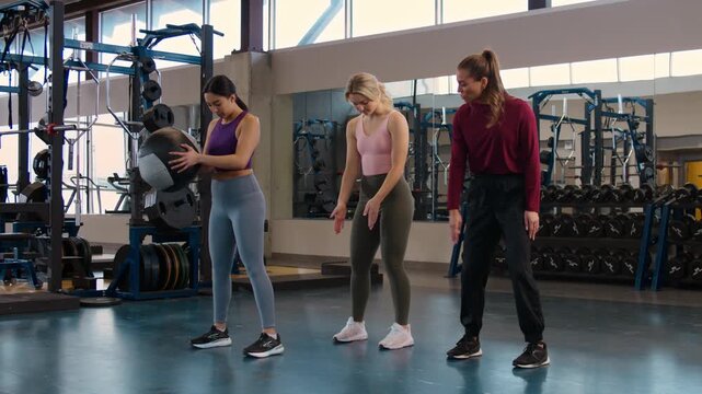 Instructor works with two female students who are learning to become certified personal trainers in a modern gym setting.