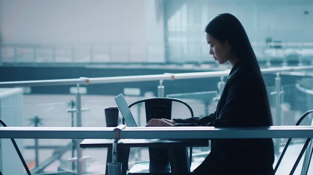 A focused Asian woman in formal attire is working on her laptop at a stylish office desk. The contemporary workspace features glass elements and a calm atmosphere, ideal for productivity.
