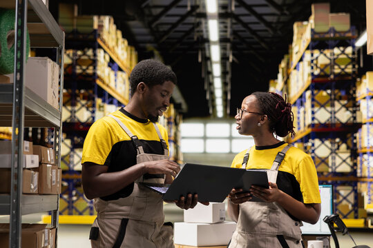 Black team members handling package data on laptop for order processing in logistics hub, verifying pallets shipping labels for distribution protocols. Efficient e-commerce warehouse.