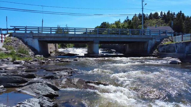 Low-level flight over a cascading river passing under a small rural concrete road bridge to its central pillar. Riviere-Blanche, Saint-Ulric-de-Matane, Quebec, Canada, 2025.