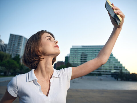 A confident woman holds a smartphone high, posing for a selfie on a sunny beach with modern buildings in the background, creating a cheerful, relaxed and upbeat outdoor mood. - Powered by Adobe