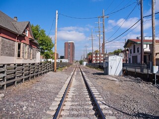 Perspective view of train tracks running through an urban neighborhood, lined with weathered wooden fences, historic buildings, and utility poles. 