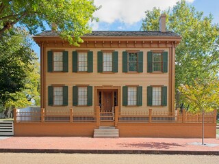Front elevation of the preserved Lincoln Home with yellow siding, green shutters, and classic wooden fence, bathed in soft daylight beneath leafy trees. 
