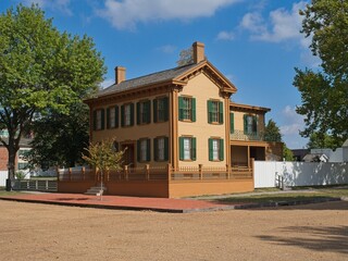 Historic yellow frame house with green shutters and wrap-around porch sits amidst trees and white picket fencing under a partly cloudy sky.  Abraham Lincoln lived here.